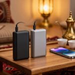 باور بانك 20000 مللي أمبير أسود ورمادي بكابلات USB-C مدمجة بجانب جوال ودلة نحاسية وفناجين وتمر على صينية خشبية فوق سجادة سدو في مجلس سعودي دافئ